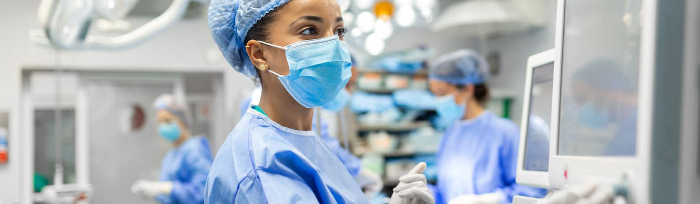 A nurse in blue scrubs and a surgical cap intently monitors data on a medical screen, operating the equipment with gloved hands. In the background, colleagues in similar attire are focused on their tasks in a bright operating room, filled with surgical lights and advanced equipment, reflecting a sterile and professional atmosphere.