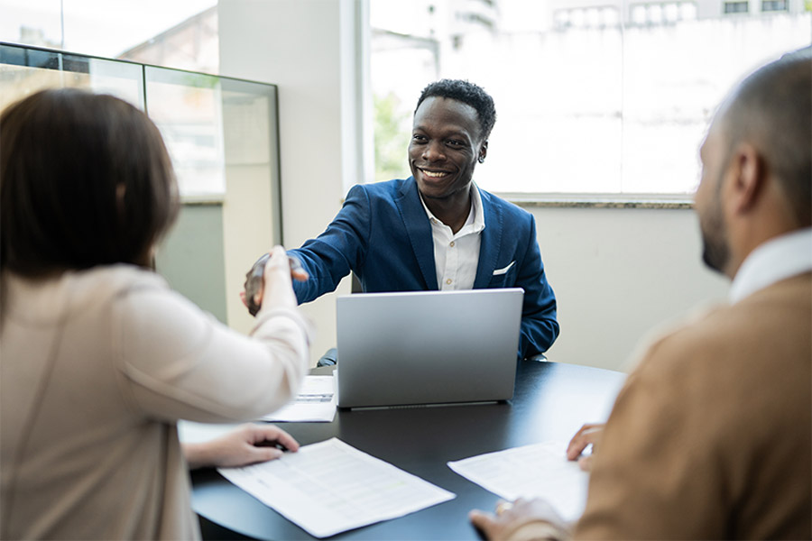 The image displays a professional consultation setting where a smiling man in a blue suit extends a handshake across a table, likely greeting or concluding a meeting. He sits behind a laptop, indicating a business or consultation environment. Papers are visible on the table, suggesting that a formal discussion or review is taking place. The individuals facing him are partially shown, with the focus on their engagement in the interaction.