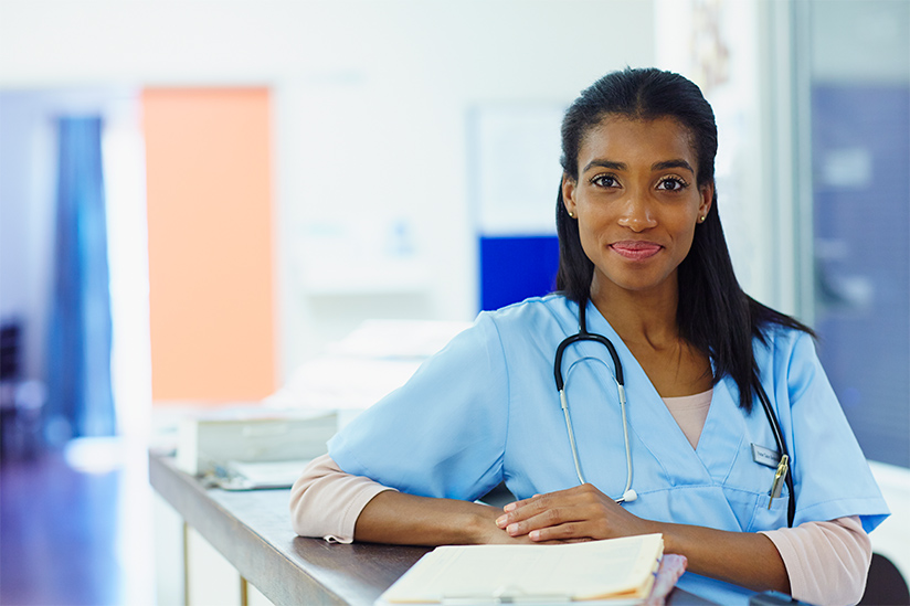 A confident female nurse wearing light blue scrubs is standing and leaning her arm on a hospital countertop. She is slightly smiling and looking straight ahead.