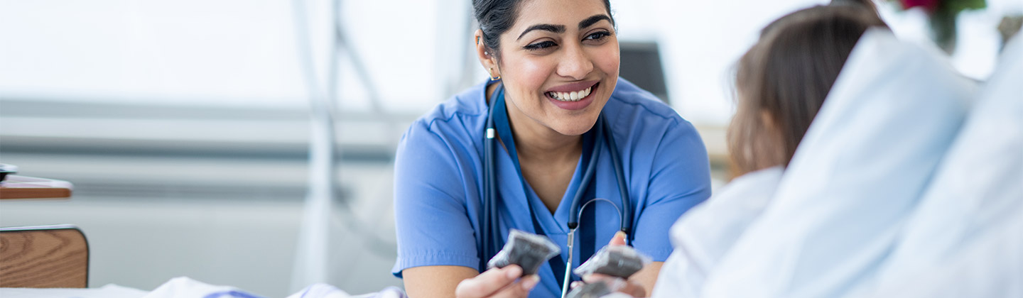 A female nurse of Middle Eastern decent sits at the edge of a hospital bed as she checks in on her young patient. She is wearing blue scrubs and is attempting to cheer the young girl up.