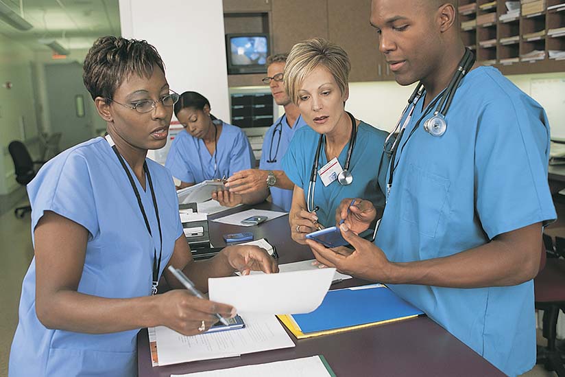 Three nurses are standing at the nurse’s station in a hospital and having a discussion. There is printed information on the desk, and they are looking at information on a small screen, possibly a pager.