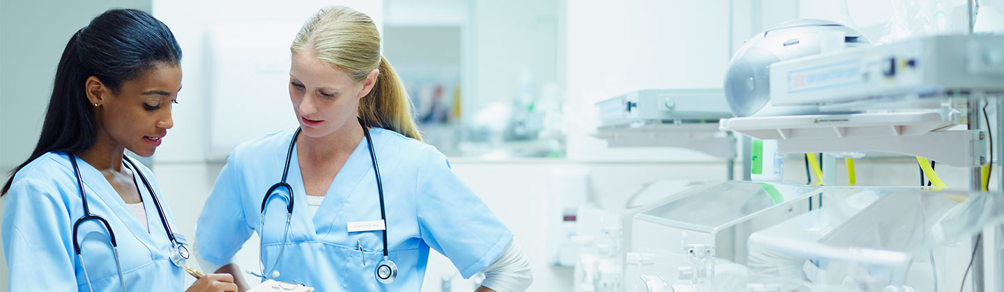 Two female nurses wearing light blue scrubs are standing in a neo-natal intensive care unit and looking at a clipboard while discussing a change of shift report. There are incubators and monitoring equipment lined up against a wall.