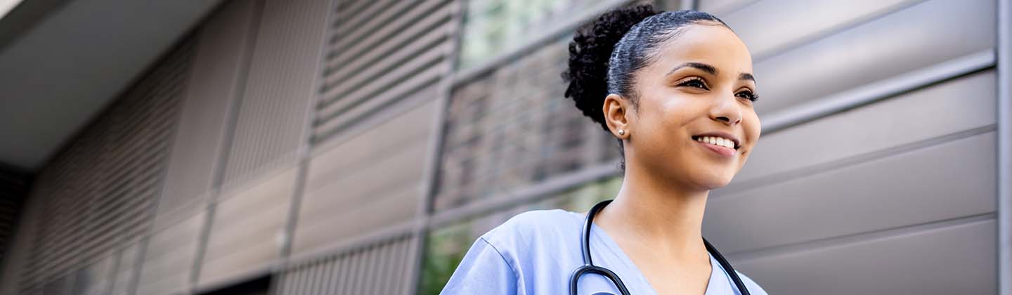 A smiling female nurse dressed in blue scrubs is walking outside of an office building.