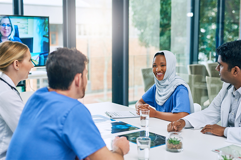 A group of culturally diverse nurses are seated at a conference table attending a meeting. They are smiling and engaged in conversation.