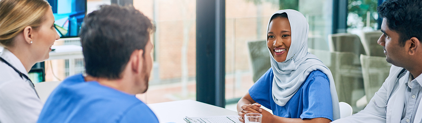 A group of culturally diverse nurses are seated at a conference table attending a meeting. They are smiling and engaged in conversation.