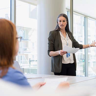 A female Director of Nursing stands in front of a conference table and is looking at a group of seated employees while giving a presentation and pointing to some information on a whiteboard behind her.