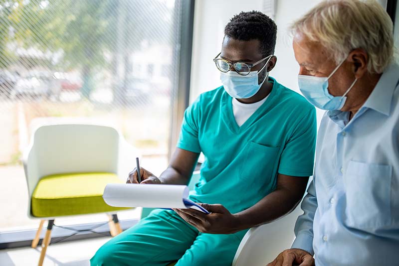 An LPN in green scrubs is sitting beside an elderly patient in a waiting area, both wearing masks. The nurse is holding a clipboard and attentively discussing the information with the patient, who is gesturing towards the clipboard and appears to be actively involved in the conversation.