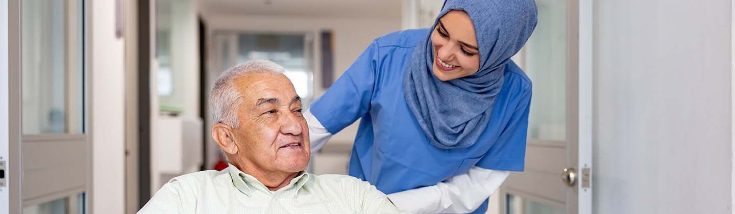 A nurse in blue scrubs and a hijab is gently pushing an elderly gentleman in a wheelchair through a hospital corridor. The nurse is leaning forward slightly, smiling and engaging with the patient, who looks relaxed and content.