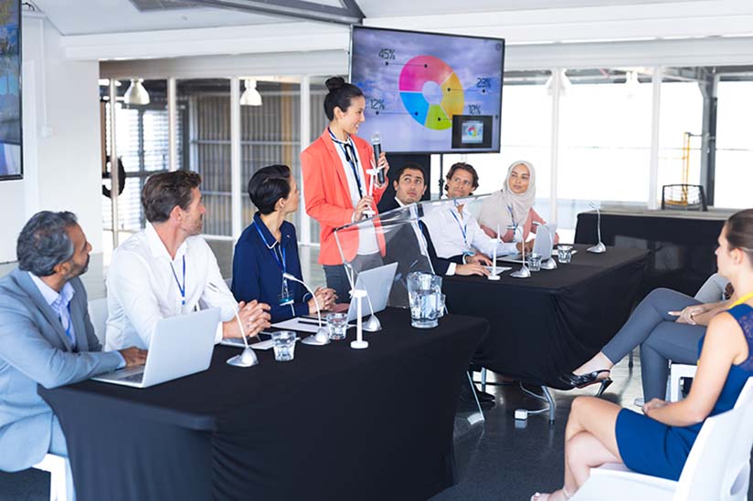 A professional meeting in a contemporary office setting with a diverse group of participants seated around an oval table. A woman in an orange blazer stands at the head of the table presenting a colorful pie chart on a monitor. The attendees, dressed in business attire, appear engaged, some taking notes on their laptops.