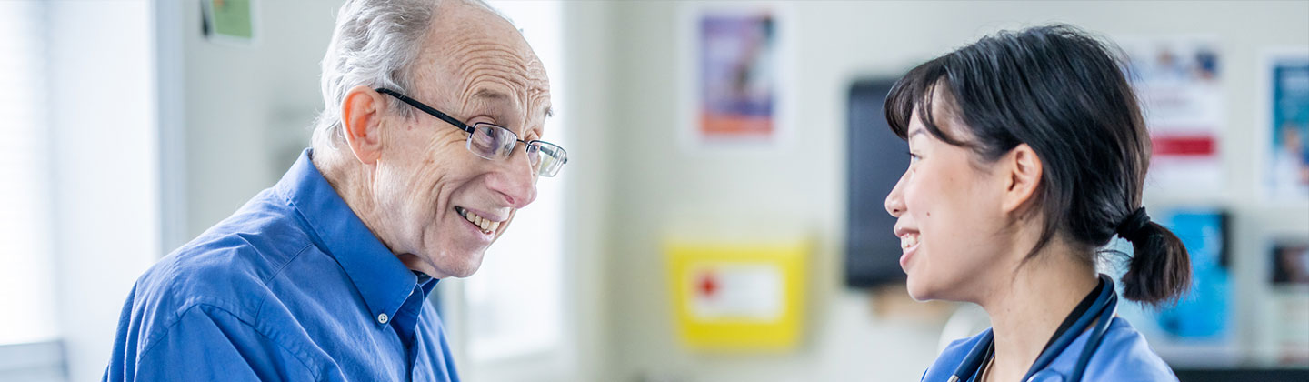 A nurse in a blue scrub top and badge provides coaching to a smiling elderly man in a blue shirt, holding a tablet in a clinic office, demonstrating a patient-centered approach in nursing.