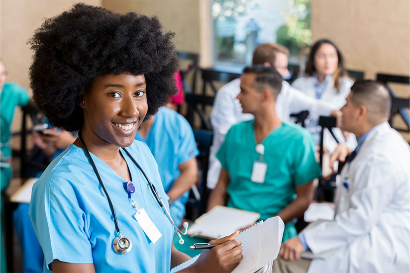 Smiling young nurse with an afro holds a clipboard in a classroom with fellow nurses chatting behind.