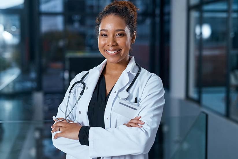 A confident African American nurse wearing scrubs and a while lab coat stands with her arms crossed in a darkened hospital hallway. She is smiling and looking straight ahead.