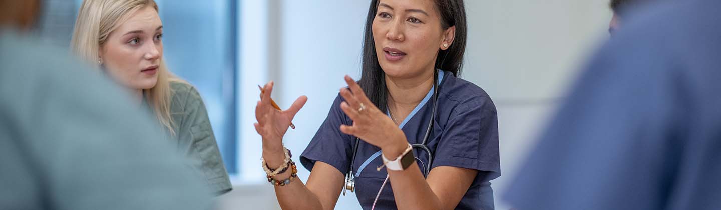 A female nursing leader is seated at a table and meeting with her staff, who are also seated. She is using her hands to emphasize a point, and her staff is looking at her and listening intently.