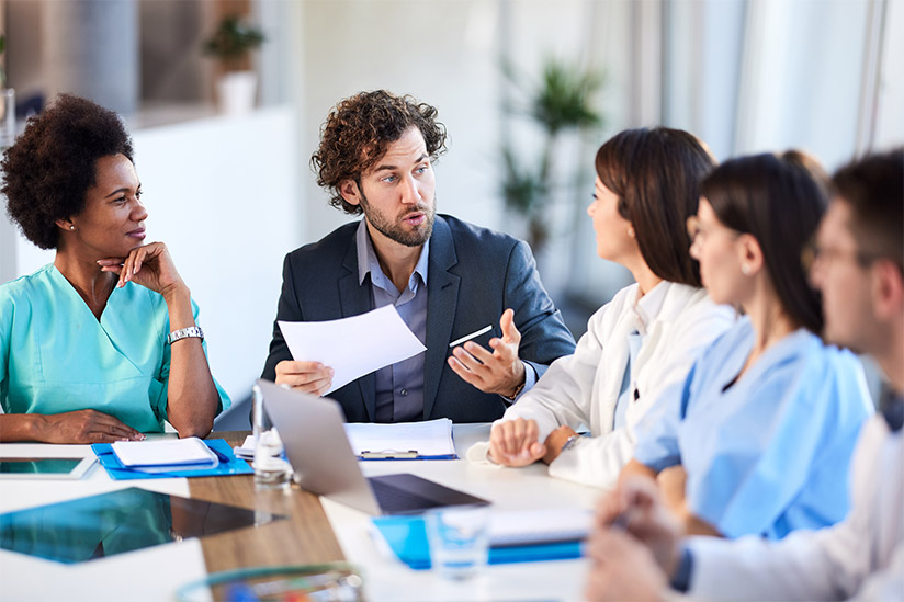 A Male Chief Nursing Executive is seated at a table with four nurse managers. They are engaged in a serious discussion of professional practice issues.