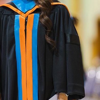 %20Image%20of%20a%20doctoral%20student%20standing%20while%20wearing%20their%20black%20graduation%20gown%20which%20has%20an%20orange%20and%20blue%20hood%20and%20front%20detail.%20Only%20their%20body%20is%20visible.%20