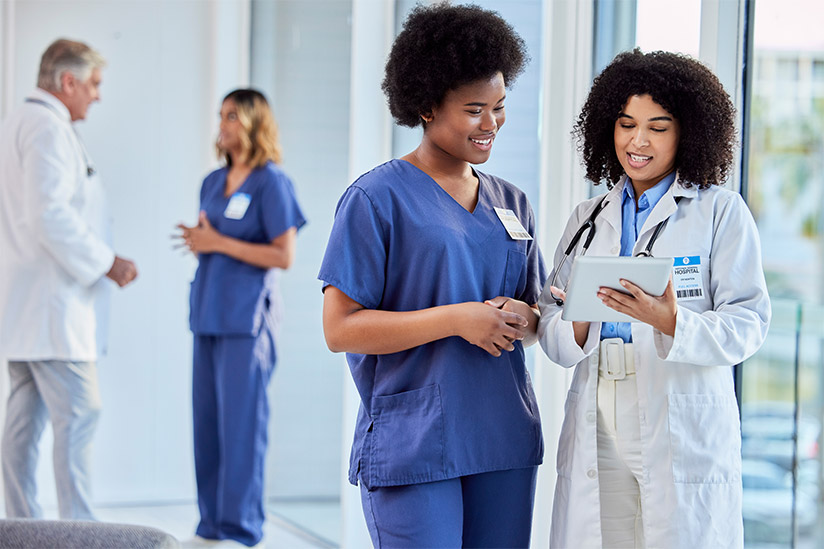 Two nurses, one holding a tablet, discuss patient care, exemplifying the use of informatics in nursing to improve healthcare delivery. Background shows peers in discussion.