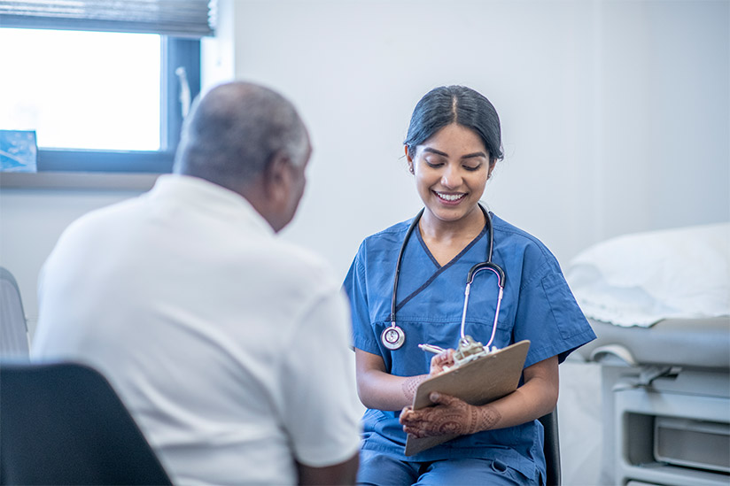 A cheerful nurse in blue scrubs takes notes on a clipboard while engaging with an elderly patient in a clinic room, signifying a caring and professional nurse-patient interaction.